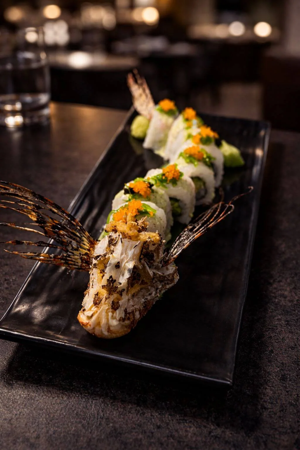 Sushi roll topped with orange roe and scallion, presented with a fried whole fish head and fins